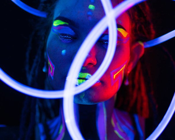 Porträt eines Mädchens mit glühenden Röhren in Neon-UF-Licht. Model Girl mit Dreadlocks und fluoreszierendem, kreativem psychedelischem Make-Up, Kunstdesign der weiblichen Disco-Tänzerin Model in UV, buntes, abstraktes Make-up - Foto, Bild