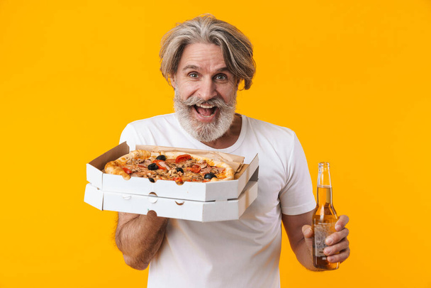 Photo of surprised emotional senior grey-haired bearded man posing isolated over yellow wall background holding pizza drinking beer. - Foto, Bild