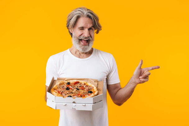 Image of happy optimistic emotional senior grey-haired bearded man posing isolated over yellow wall background holding pizza pointing aside. - Фото, изображение