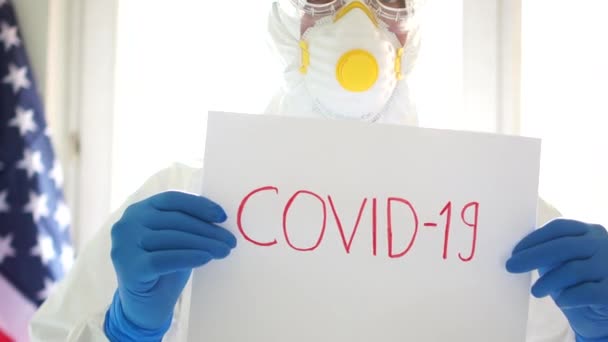 ABD 'de Covid19 Coronavirüs salgını. Koruyucu takım elbiseli bir adamın Covid19 yazan bir tabela tutarken çekilmiş yakın bir portresi. Karantina ve kendini izole etme - Video, Çekim