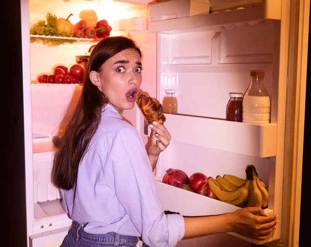 Young woman eating junk croissant near fridge in night - Photo, image