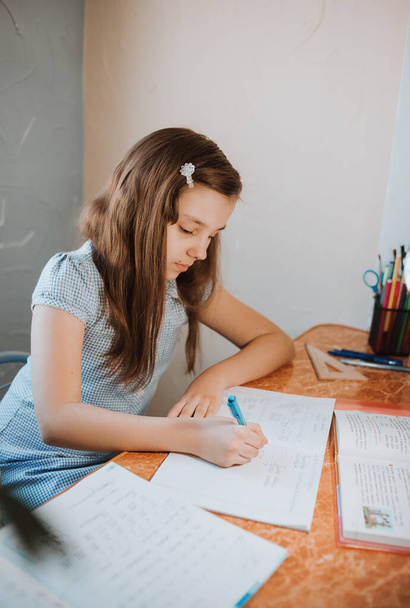 girl writes homework in a notebook at home during CoVid-19 quarantine, distance learning online with a laptop, a child doing homework for school. Children stayed at home. Girl writes in Cyrillic. - Photo, Image