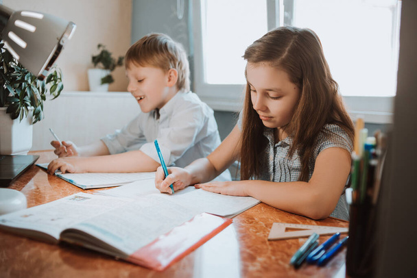The girl and boy writing them homework in the copybook at home during CoVid-19 quarantine, distance learning online with a laptop, a child doing homework for school. Children stayed at home. - Фото, изображение