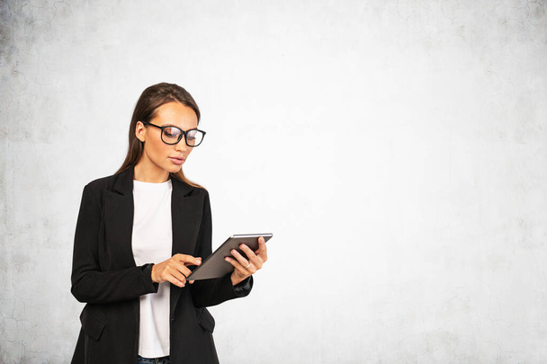 Portrait of serious young European businesswoman with glasses using tablet computer. Concrete background. Concept of internet, communication and education. Mock up - Fotografie, Obrázek