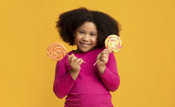 Hübsches kleines schwarzes Mädchen posiert mit zwei bunten Lutschern in den Händen - Foto, Bild