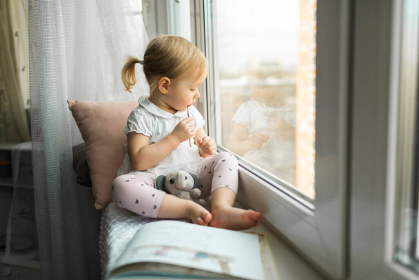 Little girl sitting on the windowsill of the house, self-isolation on quarantine at home - Foto, afbeelding