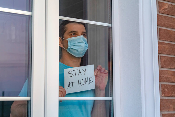 Молодой человек в медицинской маске стоит дома за окном и показывает табличку с надписью "Оставайтесь дома". Призывы к самоизоляции из-за пандемии и риска коронавирусной инфекции COVID-19
 - Фото, изображение