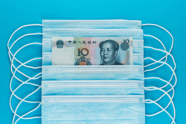 10 juanów w stercie masek medycznych. Wzrost cen masek medycznych. COVID-19 koronawirus w Chinach. Niedobór masek do twarzy. Niebieskie maski medyczne i pieniądze. Widok z góry, zbliżenie - Zdjęcie, obraz