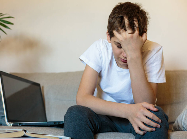 Cute upset boy in white t shirt sitting on the couch in the living room next to laptop and study. Stressed, tired kid. Home education, self education by kids. - Zdjęcie, obraz