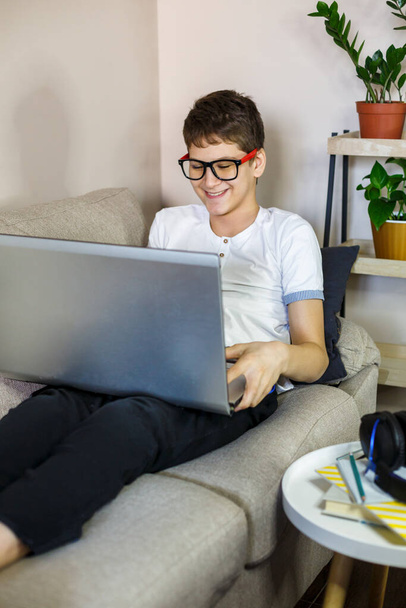 Χαριτωμένο νεαρό αγόρι με γυαλιά και λευκό t-shirt κάθεται στον καναπέ στο σαλόνι με φορητό υπολογιστή και μελέτη. Διδασκαλία κατ 'οίκον, αυτομόρφωση, εξ αποστάσεως εκπαίδευση από παιδιά. - Φωτογραφία, εικόνα