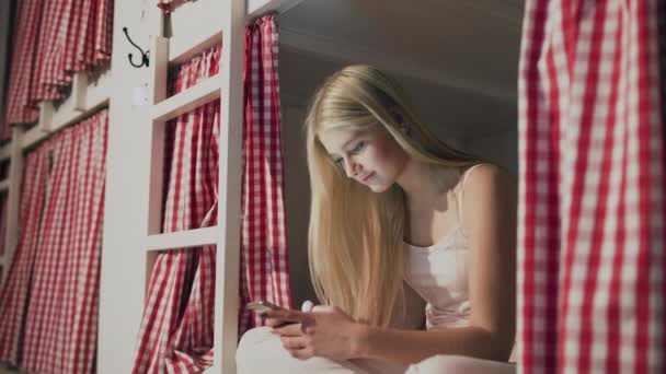 Jonge aantrekkelijke vrouw zitten op het bed in het hostel kamer en met behulp van smartphone - Video