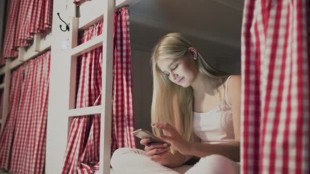 Mujer atractiva joven sentado en la cama en la habitación del albergue y el uso de teléfono inteligente - Metraje, vídeo