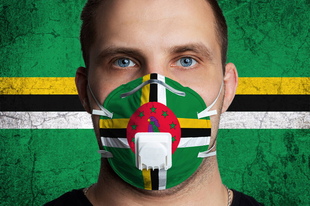 Young man with sore eyes in a medical mask painted in the colors of the national flag of Dominica. Coronovirus disease  COVID-19 concept.  Man is afraid of getting the flu - Photo, Image