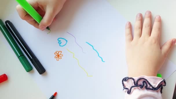 Egy kisgyerek kezei közelről. Egy gyerek zöld ceruzával rajzolja a virágokat az asztalon lévő fehér papírra.Top view.The koncepció egy boldog hétvégére, élvezi a művészetet otthon. - Felvétel, videó