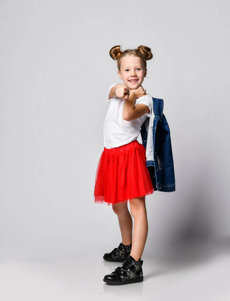 Frolic smiling blonde kid girl with funny buns in red skirt and white t-shirt is pointing finger at us - Photo, image