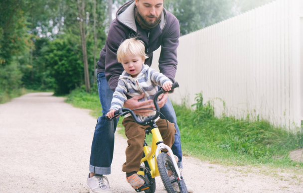 Vader leert zijn kleine kind fietsen in het voorjaarspark, vangt jongen als hij valt. Gelukkige familiemomenten. Samen tijd doorbrengen met vader en zoon. Candid lifestyle imago. - Foto, afbeelding