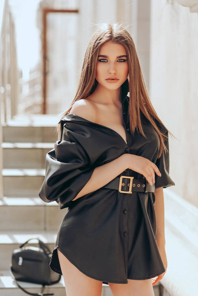 fashion outdoor photo of beautiful woman with dark hair in elegant black leather dress and accessories posing in the city street - Photo, Image