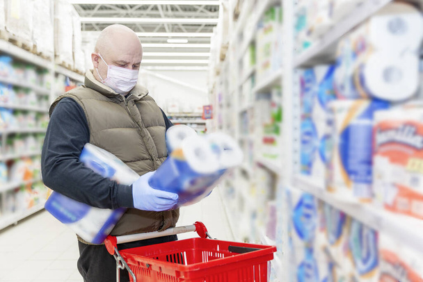 Ένας φαλακρός ενήλικας με ιατρική μάσκα και γάντια διαλέγει χαρτί τουαλέτας σε σούπερ μάρκετ. Θολή. Ο τρόπος της αυτο-απομόνωσης κατά την περίοδο του coronavirus pardemia.  - Φωτογραφία, εικόνα