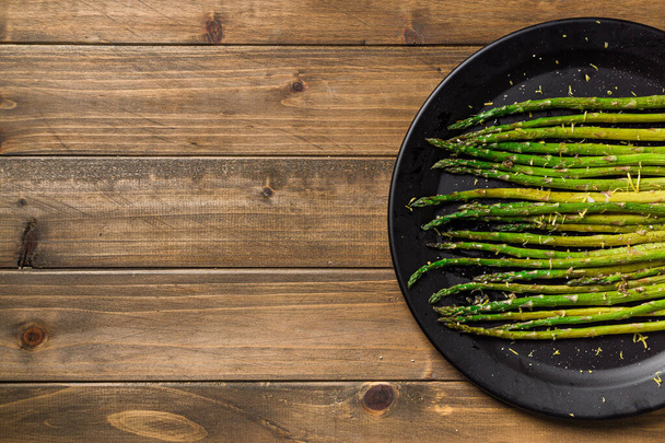 Asparagus on wooden background. Eating healthy food is easy with this homemade vegan dish; roasted asparagus with lemon zest on top. The veggies are served on black plate with wooden table background. - Photo, Image