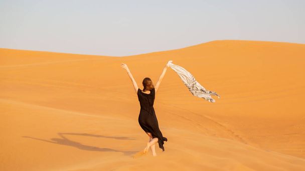 A girl walks on the sand in a deserted desert. A woman in a black dress with a cloth in her hands in a strong wind alone in the desert - Fotoğraf, Görsel