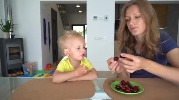 Loving mummy feeding her child boy with cherry berry at home. Mother kissing son - Felvétel, videó