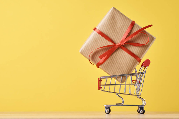 Einkaufswagen und Geschenk auf gelbem Hintergrund. Ferien- und Geburtstagskonzept - Foto, Bild