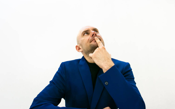 Hombre de negocios moden en chaqueta azul sostenga su dedo en la mejilla y mira hacia arriba mientras piensa. Imagen de espacio en blanco. Concepto de toma de decisiones
. - Foto, Imagen