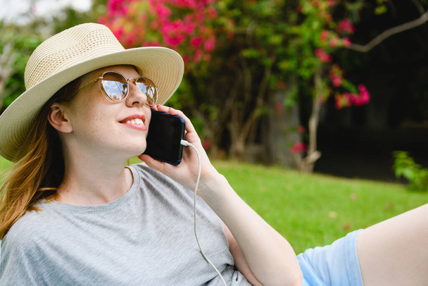 Genç bir kadının yüzü akıllı telefonu kablo TV 'de açık, şapka takıyor ve güneş gözlüğü takıyor, doğal yeşil arka planda yakın plan. - Fotoğraf, Görsel