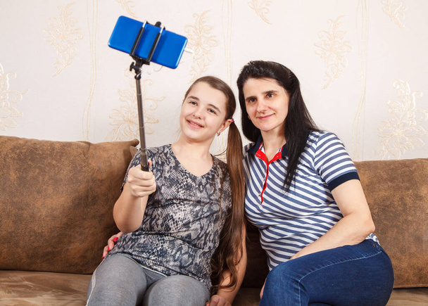 Мама и дочь делают селфи со смартфоном, сидя дома на диване. Карантин коронавируса
. - Фото, изображение