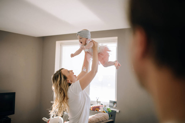 Mamá sostiene al bebé en sus brazos en la habitación luminosa de su casa. El marido mira a su esposa e hija mientras se divierten
. - Foto, imagen