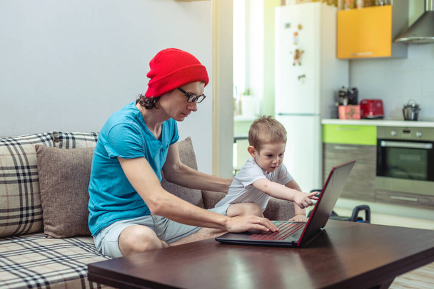 膝に子供がいる若い父親がノートパソコンで家に座っている。自宅に滞在する必要があるときの距離の仕事とオンライン教育の概念. - 写真・画像