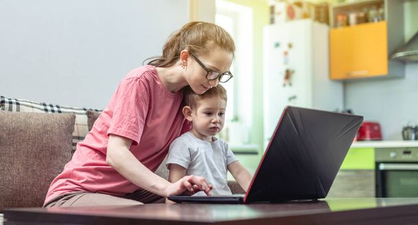 A young woman with a small child on her knees is sitting at a laptop. Concept of remote work and receiving online education from the comfort of home. - Photo, Image