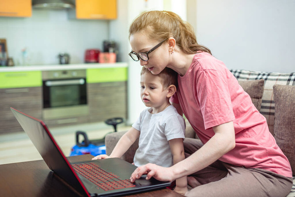 Młoda kobieta z małym dzieckiem na kolanach siedzi przy laptopie. Koncepcja pracy zdalnej i otrzymywanie edukacji online w zaciszu domu. - Zdjęcie, obraz