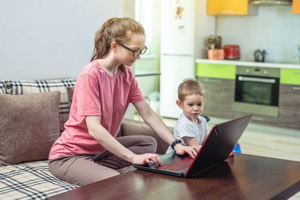 膝の上に小さな子供を持つ若い女性がノートパソコンに座っている。リモートワークの概念と自宅の快適さからオンライン教育を受ける. - 写真・画像
