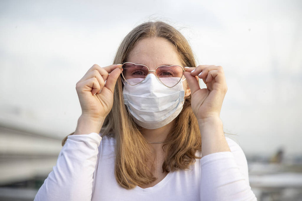 Portrait of a beautiful caucasian girl in a mask and pink glasses in the shape of hearts looks at the camera. in the background planes and the airport in blur. Coronaviirus covid19 concept - Photo, Image