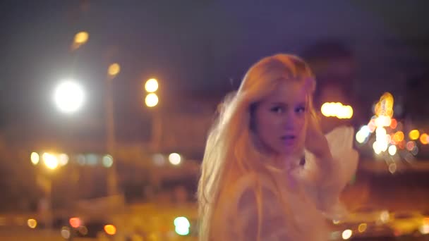 Krásná kavkazská dívka módní model procházky podél nočního města, šťastný a usmívající se. Mladá žena s make-upem v bílém stylové šaty narovnává její dlouhé vlasy a pózuje na pozadí světel - Záběry, video