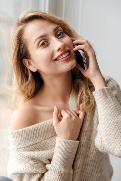 Photo of a smiling young lady indoors at home talking by mobile phone near window. - Foto, afbeelding