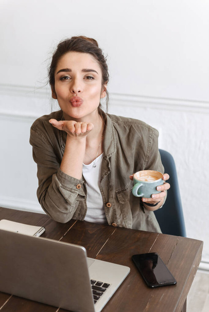 Lovely young woman using laptop computer while sitting at the cafe indoors, drinking coffee, sending kiss - Фото, изображение