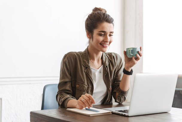 Adorável jovem mulher usando computador portátil enquanto sentado no café dentro de casa, bebendo café
 - Foto, Imagem