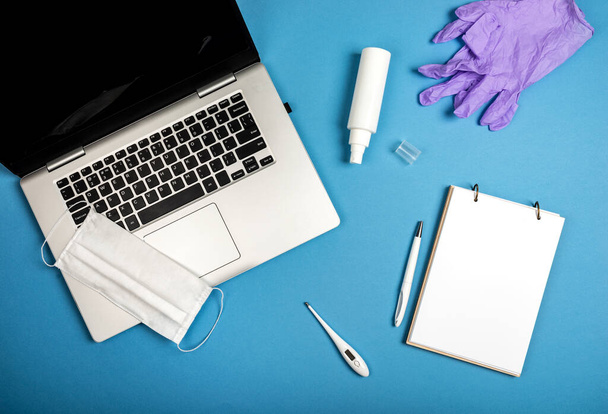 Laptop, notebook, toll, sebészeti maszk, orvosi kesztyű, kézfertőtlenítő és digitális hőmérő a kék háttérben. Online munka vagy oktatás a karantén feltételei között. Felülnézet. - Fotó, kép
