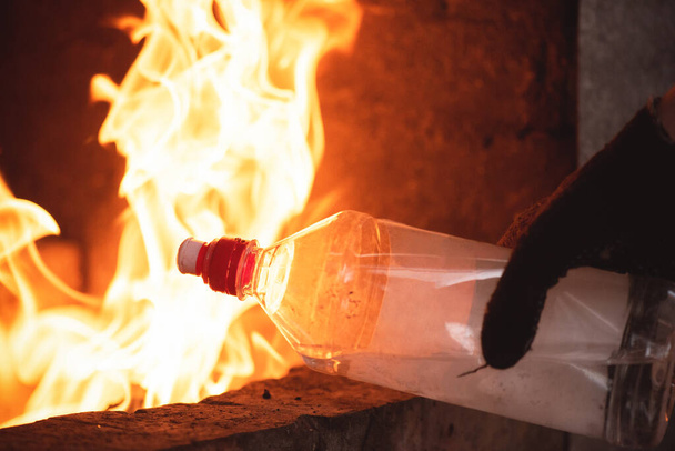 Hand in Hand mit einer Zündflüssigkeitsflasche und brennendem Feuer im Kaminhintergrund aus nächster Nähe. - Foto, Bild