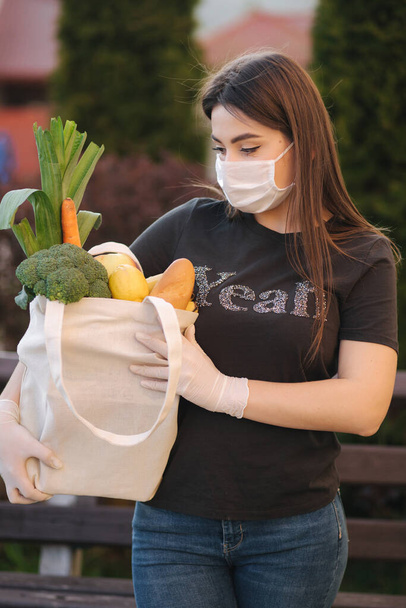 Fiatal nő maszkban és kesztyűben tartja táska friss étel, zöldség gyümölcsök és kenyér. Nő a bolt után. Védje magát. Coronavirus téma - Fotó, kép