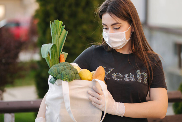 Mujer joven con máscara y guantes bolsa de mano con alimentos frescos, verduras frutas y pan. Mujer después del supermercado. Protéjase. Tema Coronavirus
 - Foto, Imagen