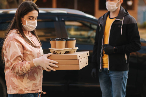 Młoda kobieta w masce ochronnej i masce medycznej przyjmuje zamówienie od dostawcy na zewnątrz. Dostawa żywności w kwarantannie. Kurier przynieść pizzę i kawę dla klienta - Zdjęcie, obraz