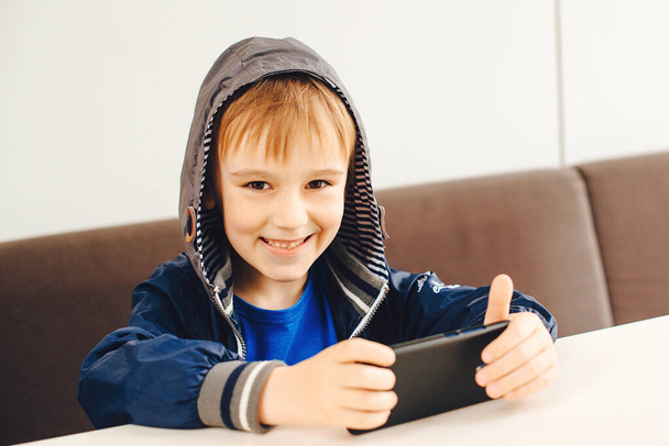 幸せな少年は携帯電話でゲームをプレイ。屋外のテラスでスマートフォンでビデオを見ている子供。現代の装置やライフスタイル。スマートフォンやインターネットを利用したスタイリッシュな男子. - 写真・画像