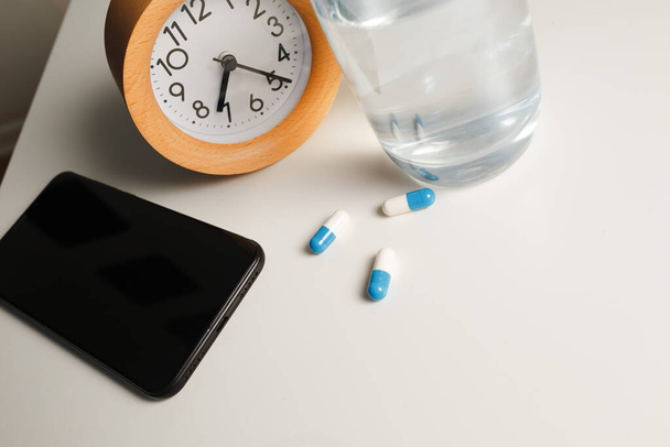 Alarm clock, glass of water, mobile phone and white pills on white table background - Photo, Image