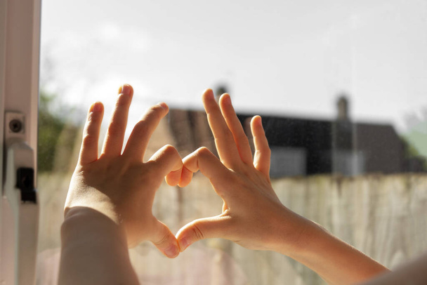 Small girl making a heart gesture with her fingers on the window. Autoisolation concept. - Foto, Imagem