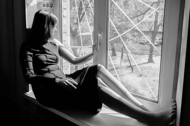 retrato preto e branco de uma linda menina de cabelos pretos sentada em um peitoril da janela em um vestido escuro apertado
 - Foto, Imagem
