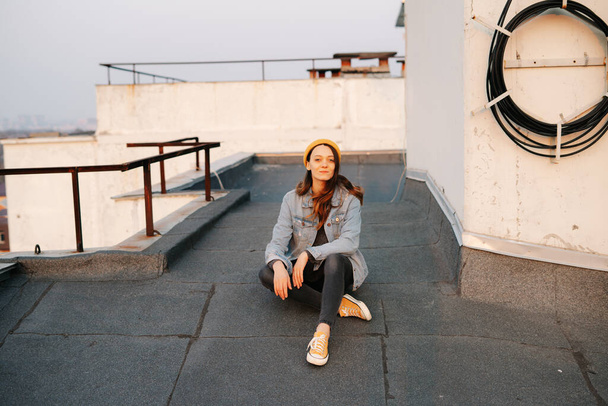 Ein hübsches europäisches, weißes Mädchen sitzt auf dem Dach eines mehrstöckigen Gebäudes in einer blauen Jeansjacke mit grauer Hose, gelbem Hut und gelben Turnschuhen.. - Foto, Bild