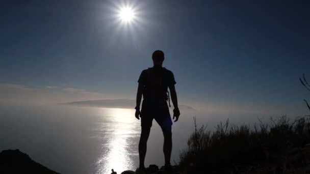 Sylwetka szczęśliwego turysty stojącego na szczycie. Zwolnij trochę. Młody inspirowany człowiek stojący na szczycie góry nad oceanem przeciwko pięknemu błękitnemu niebu. - Materiał filmowy, wideo